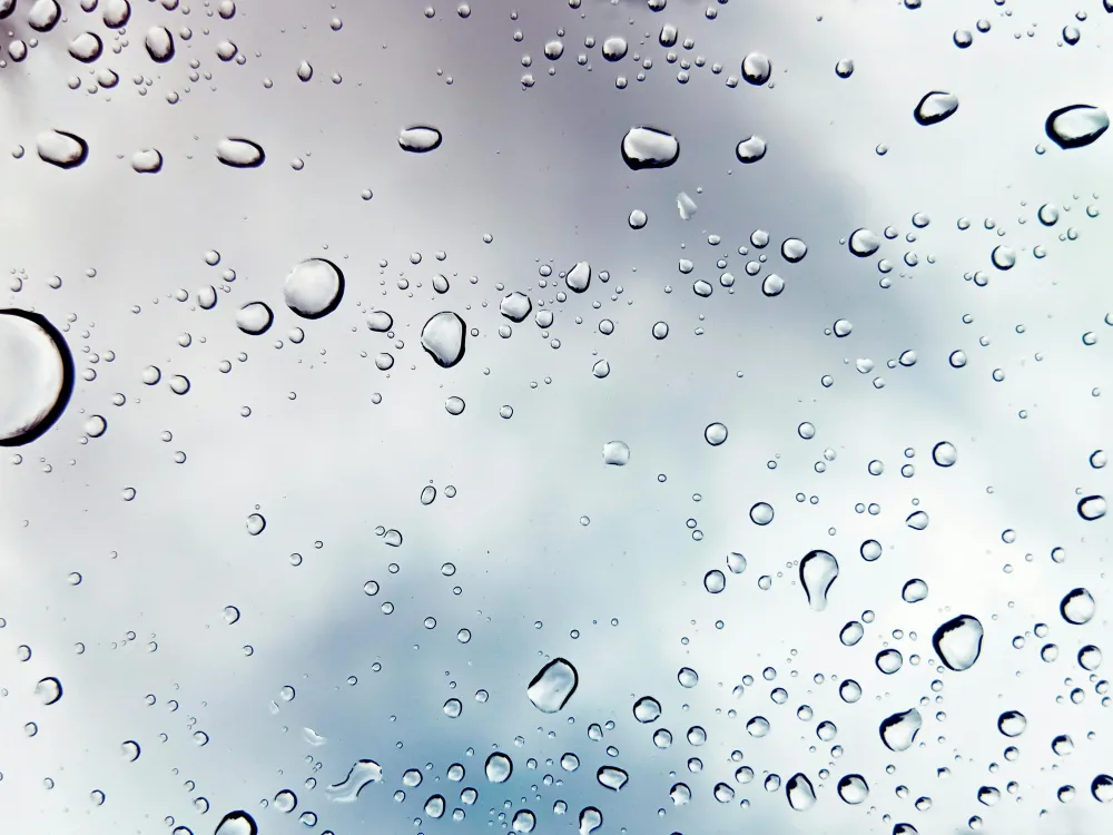 Druppels regen op een glasoppervlak met een bewolkte lucht op de achtergrond.